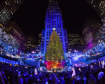 An image of the Rockefeller Center Christmas tree fully lit with buildings bathed in blue behind against the black night sky. The picture has a slight fish-eyed lens effect to focus the image on the tree.
